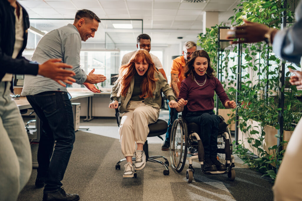 woman who uses a wheelchair and female colleague racing on chairs in an office woman who uses a wheelchair and female colleague racing on chairs in an office